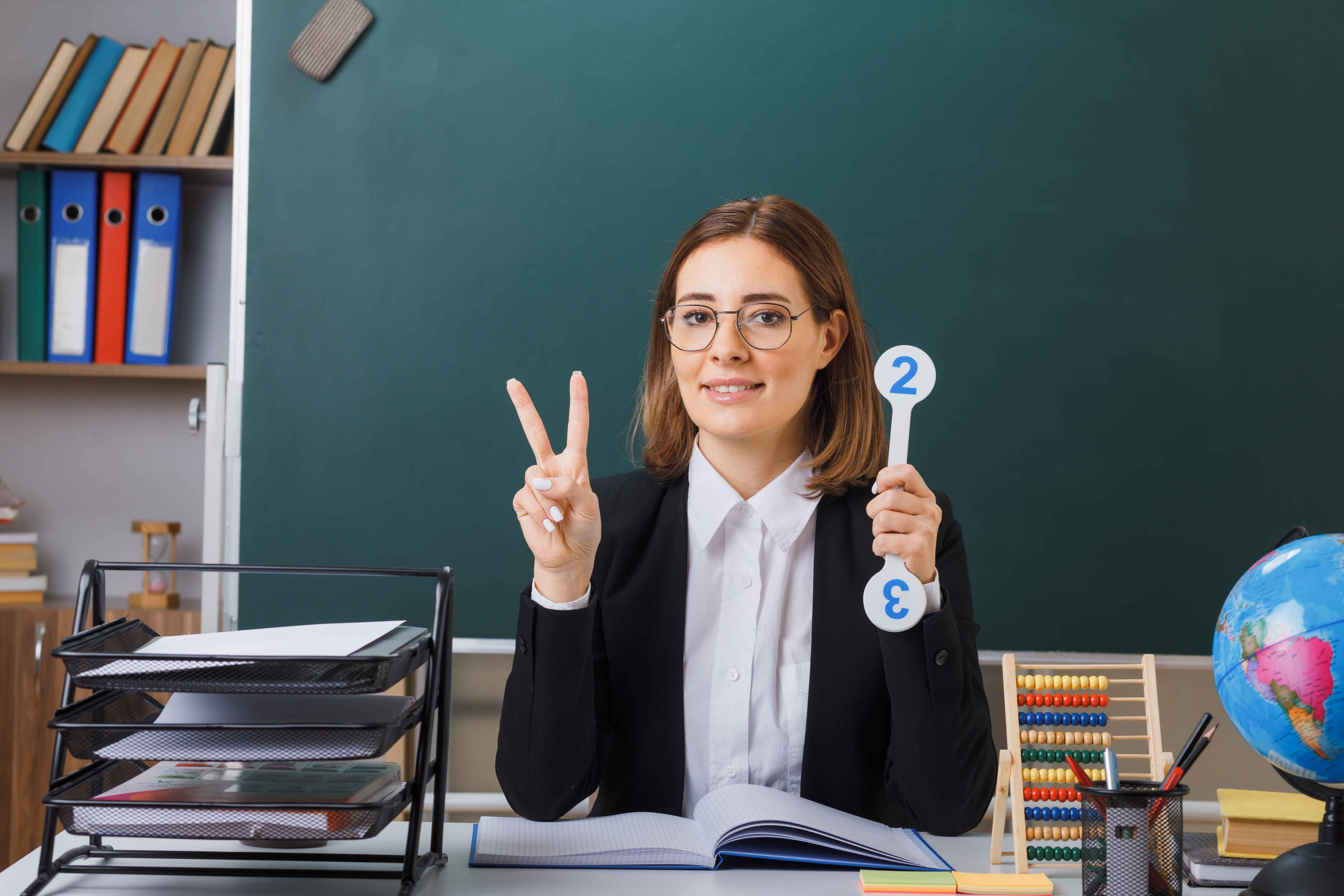 young-woman-teacher-wearing-glasses-sitting-school-desk-front-blackboard-classroom-holding-number-plates-explaining-lesson-showing-vsign-smiling-cheerfully